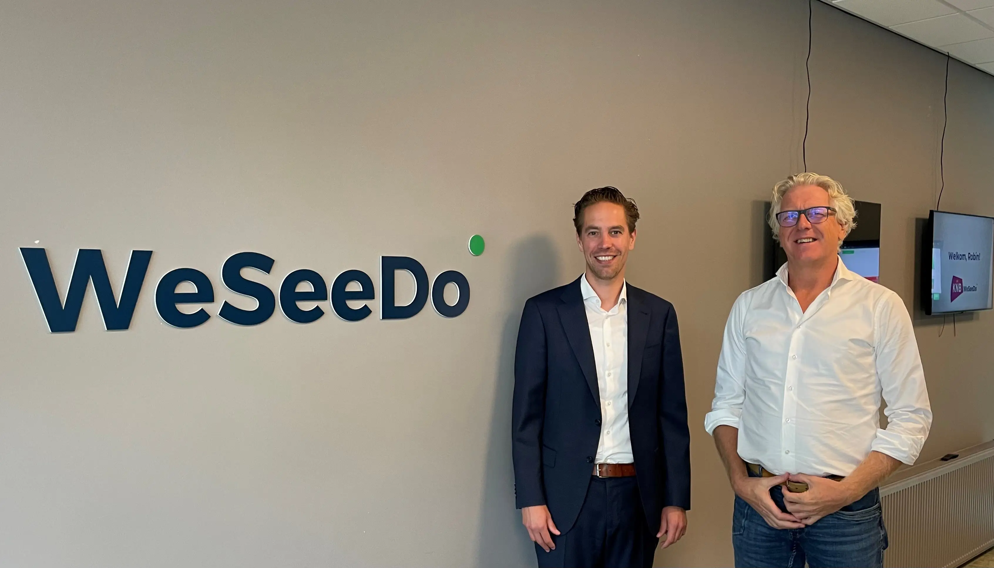 Men in suits standing in front of WeSeeDo logo in an office.