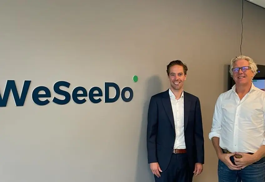 Men in suits standing in front of WeSeeDo logo in an office.