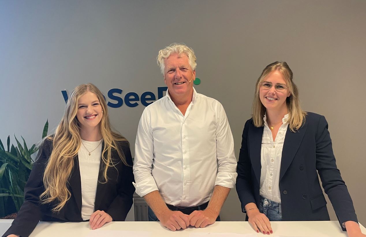 Een man en vrouw poseren glimlachend bij een balie met het logo 'WeSeeDo' op de achtergrond. De man links draagt een wit overhemd en de vrouw rechts een donkerblauwe blazer en bril.