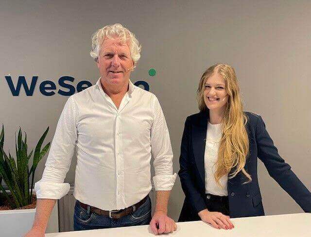 Een man en vrouw poseren glimlachend bij een balie met het logo 'WeSeeDo' op de achtergrond. De man links draagt een wit overhemd en de vrouw rechts een donkerblauwe blazer en bril.